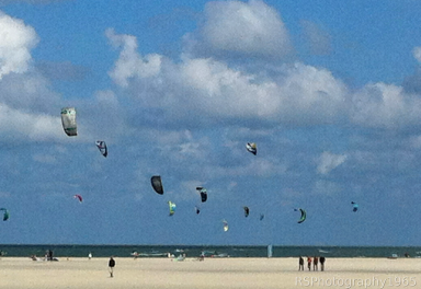 Kitesurfen in Zeeland Kitesurfers vliegen boven een zandstrand onder een blauwe lucht met wolken.